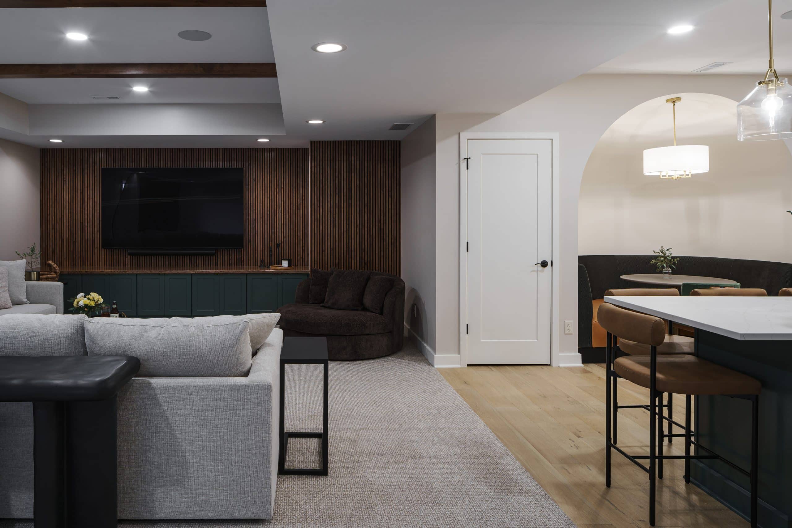 An interior view of a newly remodeled, upscale basement family room in Kansas City. The space features a cozy, modern design with a warm and inviting color palette. In the foreground is a light gray sectional sofa on a textured beige area rug. The back wall showcases a custom-built media console with dark green cabinetry and a mounted television, flanked by a striking floor-to-ceiling slatted wood accent wall. The wood paneling conceals a clever, hidden door for media storage. The room is well-lit with recessed ceiling lights and has light wood flooring transitioning from the carpeted area. The far right of the image reveals a partial view of a dining nook with a custom built-in banquette under a softly lit archway, with leather bar stools and a large kitchen island countertop in the foreground. The overall design combines rustic charm with modern functionality to create a perfect space for entertaining and family gatherings.