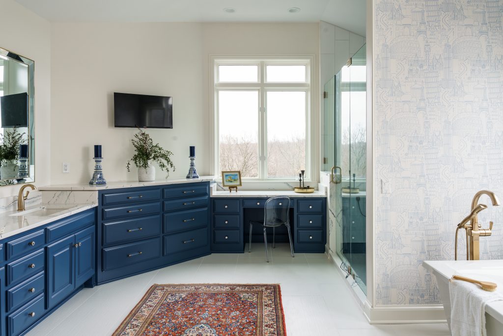 bathroom remodel that kept existing cabinets and painted them a dark blue shade with creamy white walls and counters and floor and a vintage accent rug in the middle of the room and a window over the built in vanity seatin area and the glimpse of a soaking tub with gold hardware in the corner