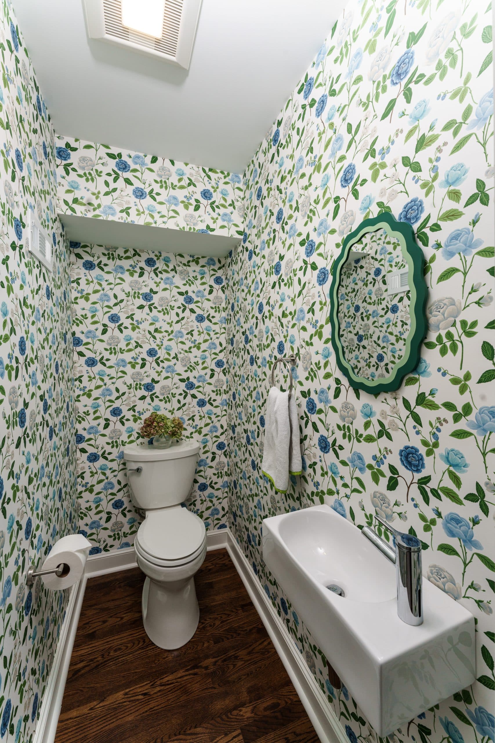 Small Powder Room Remodel with Floral Wallpaper & Floating Sink Compact powder room renovation featuring blue and green floral wallpaper, a wall-mounted floating sink, a scalloped green mirror, and dark hardwood floors.