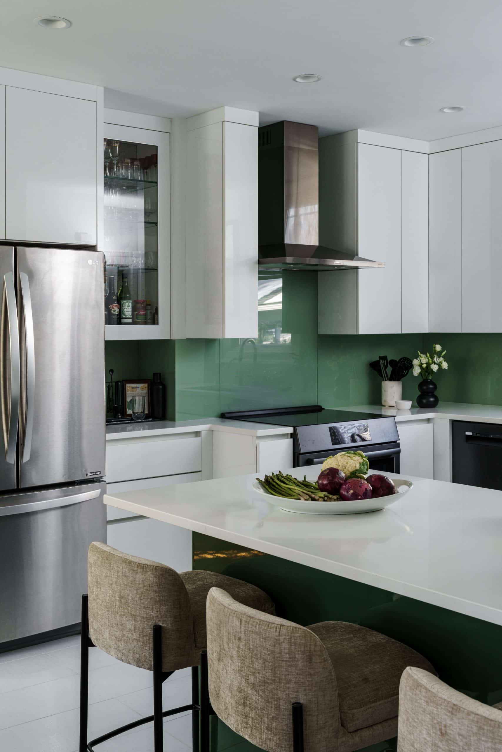 Close-up of a sleek kitchen workspace featuring a modern range hood, green glass backsplash, and custom high-gloss cabinetry. The white island with tan barstools adds to the high-design, European feel of this Brookside remodel.