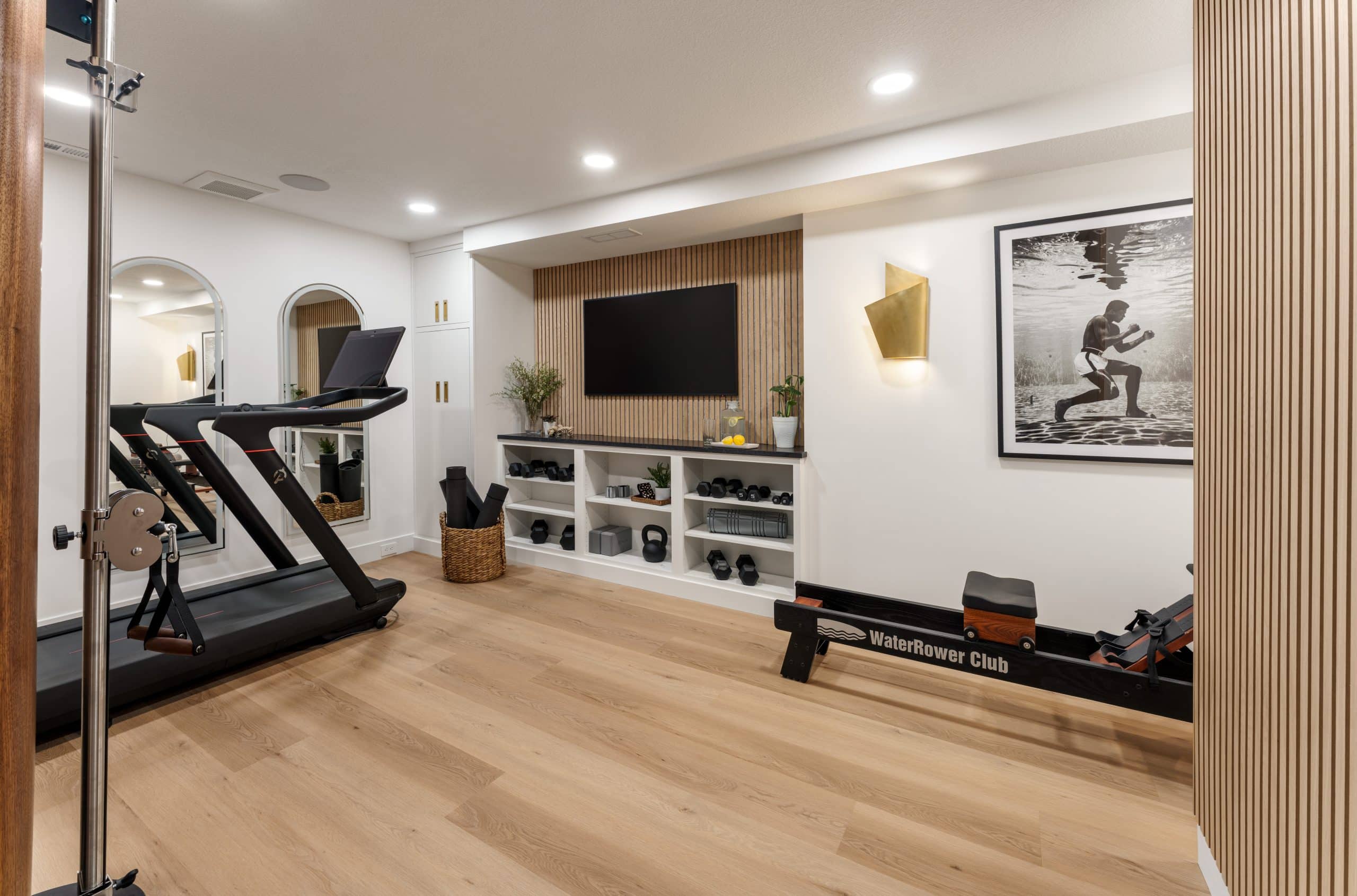 Creamy white walls and light wood flooring define this Cedar Creek basement gym remodel in Olathe, featuring a boutique fitness studio design with slat wood accent walls and black and gold finishes.