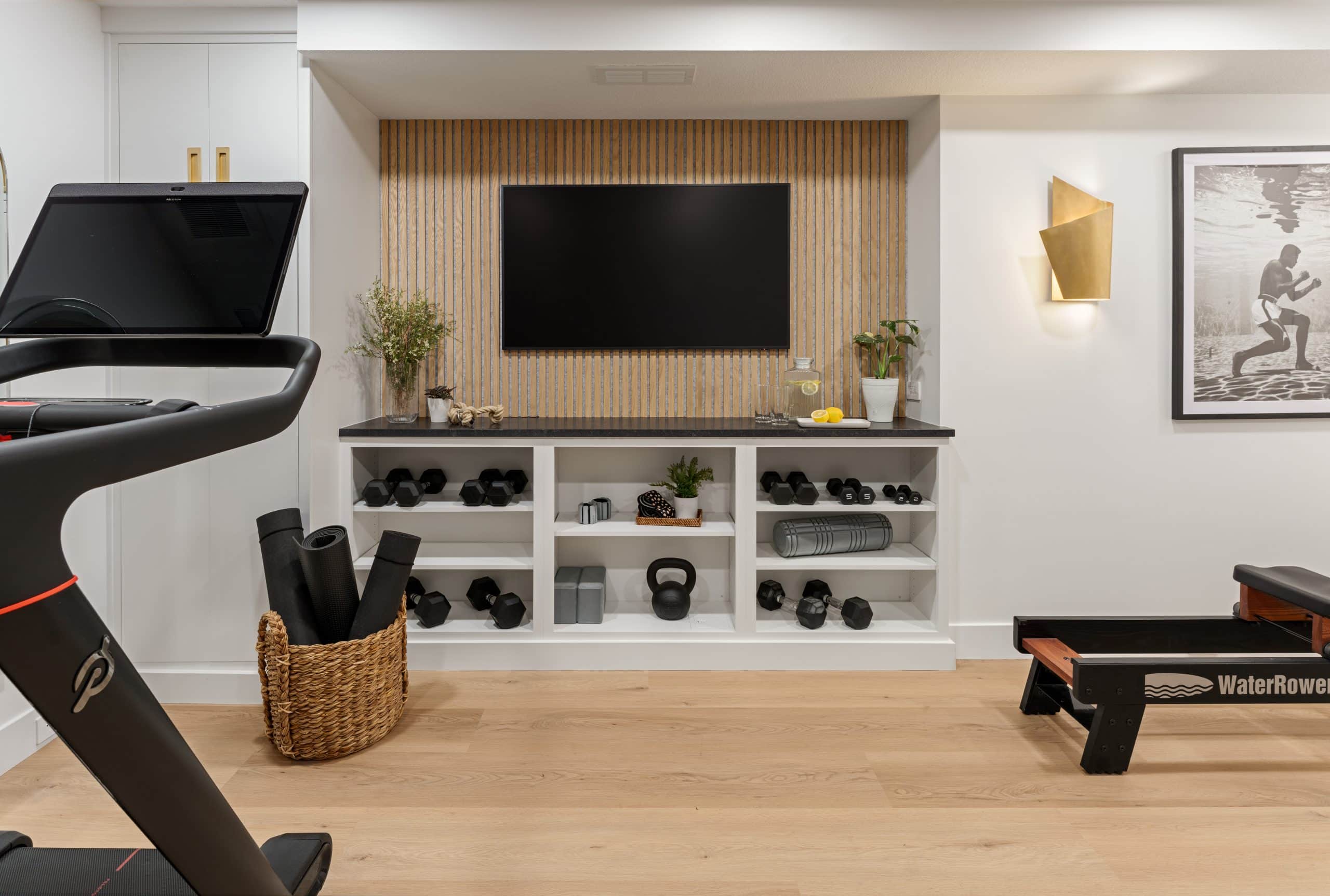 Creamy white walls and light wood flooring define this Cedar Creek basement gym remodel in Olathe, featuring a boutique fitness studio design with slat wood accent walls and black and gold finishes.